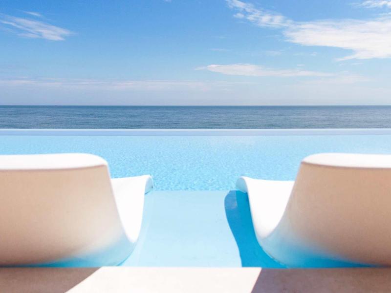 Deux chaises longues blanches au bord d'une piscine à débordement avec vue sur la mer bleue et le ciel.