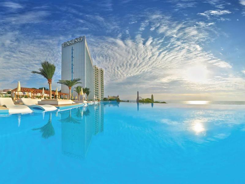 Un complexe hôtelier moderne avec une grande piscine à débordement et des chaises longues relaxantes au coucher du soleil.