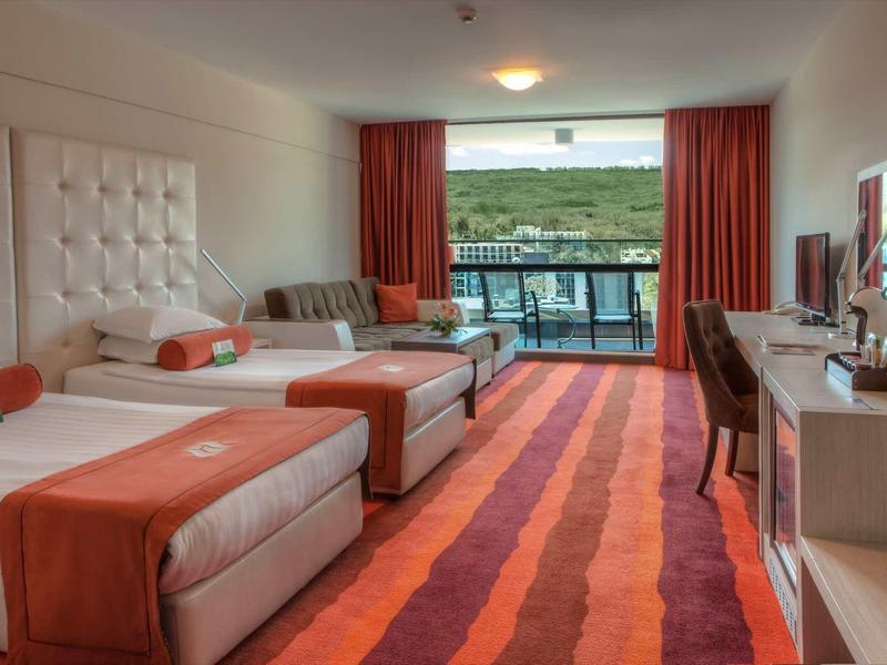 Chambre d'hôtel moderne avec deux lits simples, tapis orange et balcon donnant sur un paysage verdoyant.
