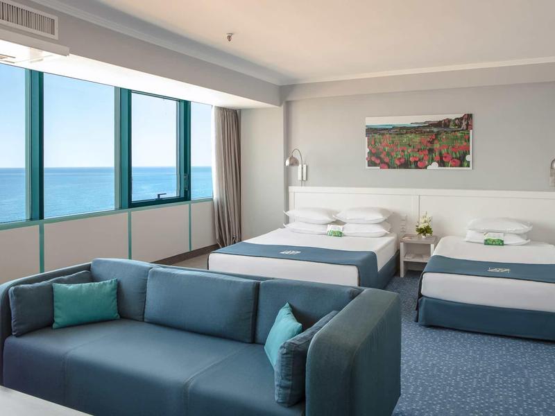 Chambre d'hôtel avec deux lits, vue sur la mer à travers de grandes fenêtres et canapé bleu au premier plan.