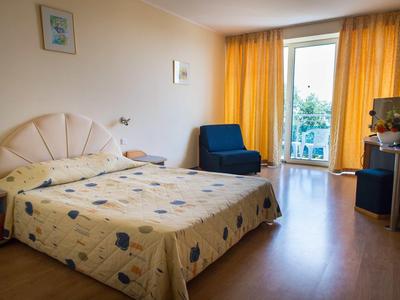 Hell beleuchtetes Schlafzimmer mit Doppelbett, gelben Vorhängen, blauem Sessel und Holzboden.