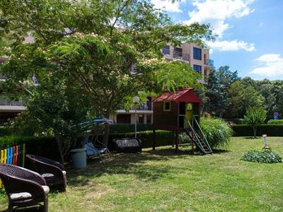 Kinderspielplatz mit Holzhaus, Schaukel, Bänken und Bäumen auf einer grünen Wiese bei sonnigem Wetter.
