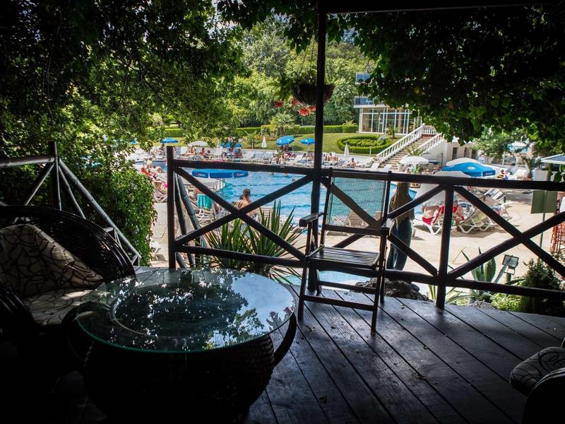 Überdachte Terrasse mit Holzboden und Gartenmöbeln, Blick auf Sonnenschirme und Bäume im Hintergrund.