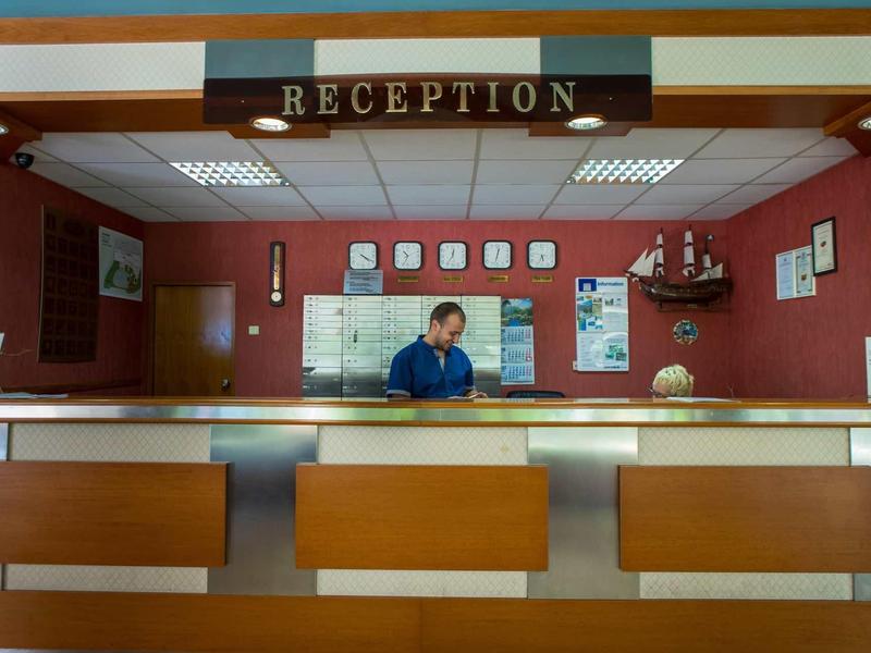 Eine Rezeption mit Holz- und Glasfront, roter Wand, Schildern und zwei Personen dahinter.