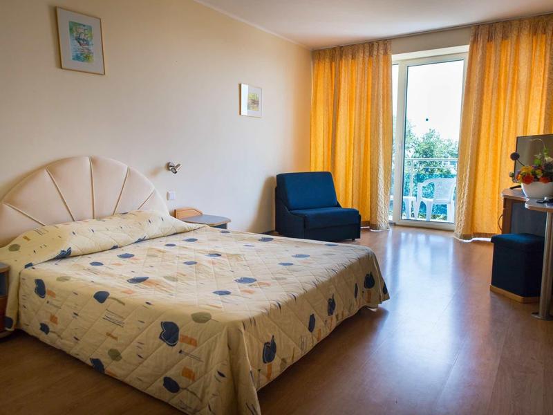 Hell beleuchtetes Schlafzimmer mit Doppelbett, gelben Vorhängen, blauem Sessel und Holzboden.