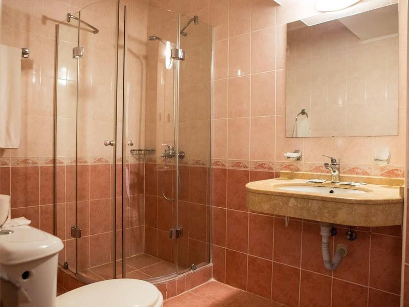 Kleines Badezimmer mit braunen Fliesen, runder Waschtisch, Spiegel und Eckdusche mit Glaswänden.