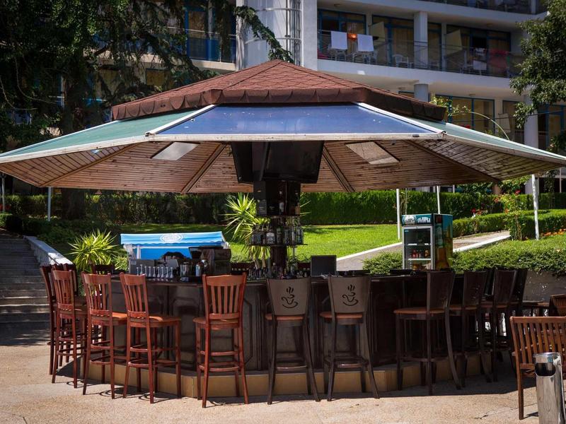 Runder Außenbarbereich mit Holzstühlen unter großem Sonnenschirm und umliegender Wohnbebauung.