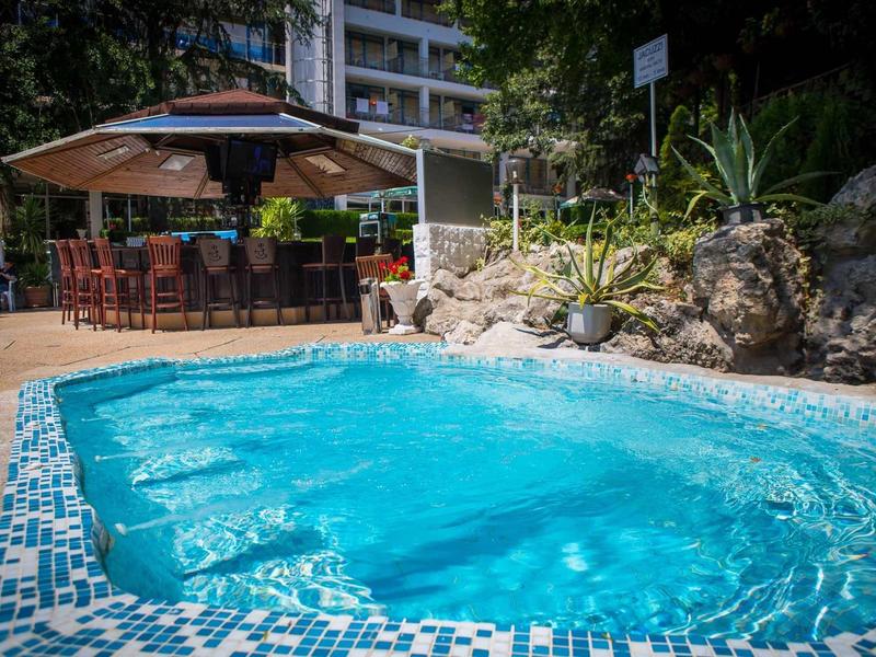 Kleiner blauer Pool mit Mosaikrand, umgeben von Felsen, Pflanzen und einer überdachten Bar im Hintergrund.