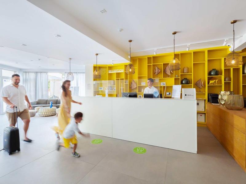 Hotelreceptie met gele planken en een familie die aankomt terwijl een medewerker klaarstaat.