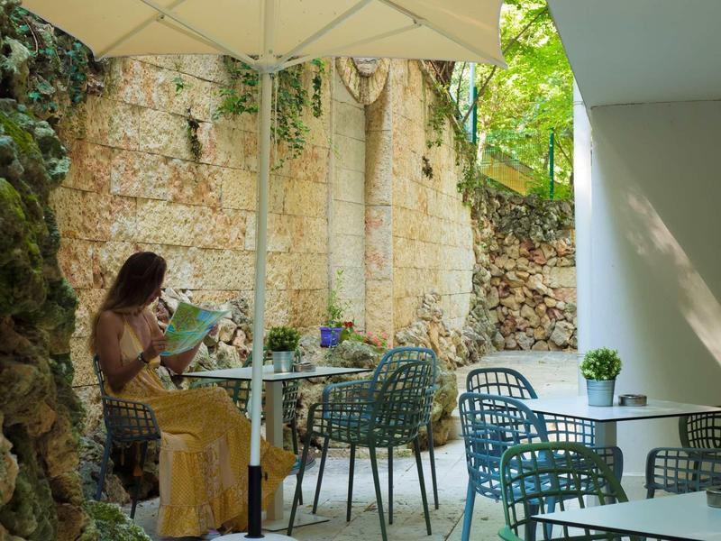 Zonnig terras met blauwe stoelen, tafels en grote parasol bij een stenen muur.