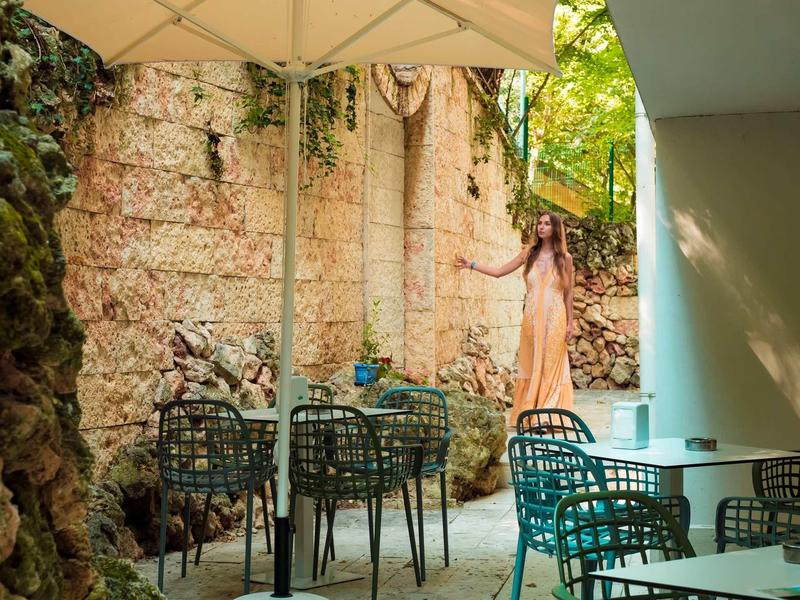 Vrouw in lange jurk staat in buitenruimte met tafels en stoelen bij stenen muur en parasol.