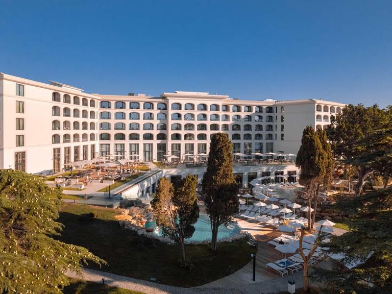 Large hotel with modern facade, pool area, and sun umbrellas outdoors under clear sky.