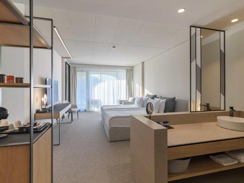 Bright hotel room with large bed, desk, shelving, and modern washbasin area.