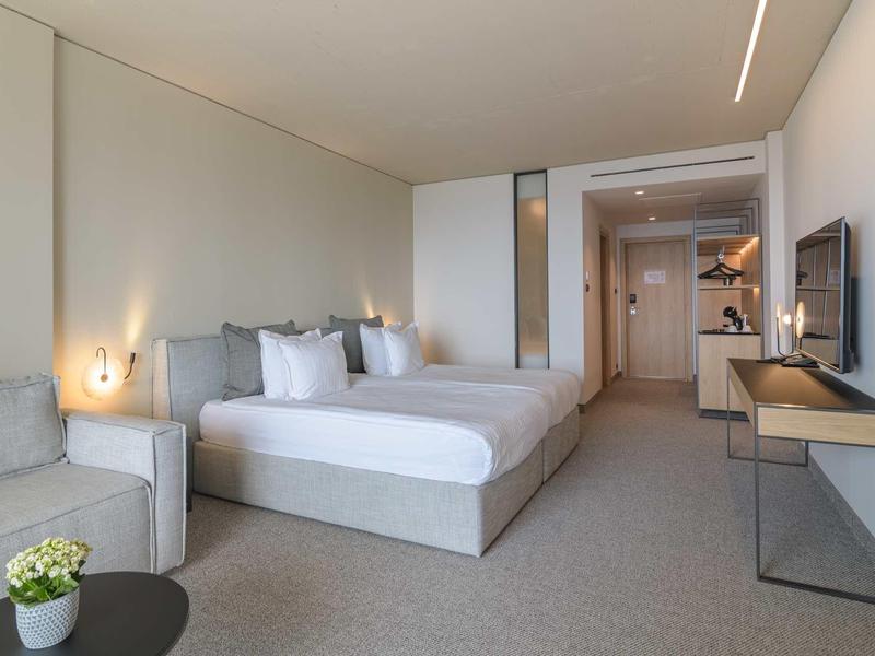 Modern hotel room with double bed, sofa, TV, and minimalist design.