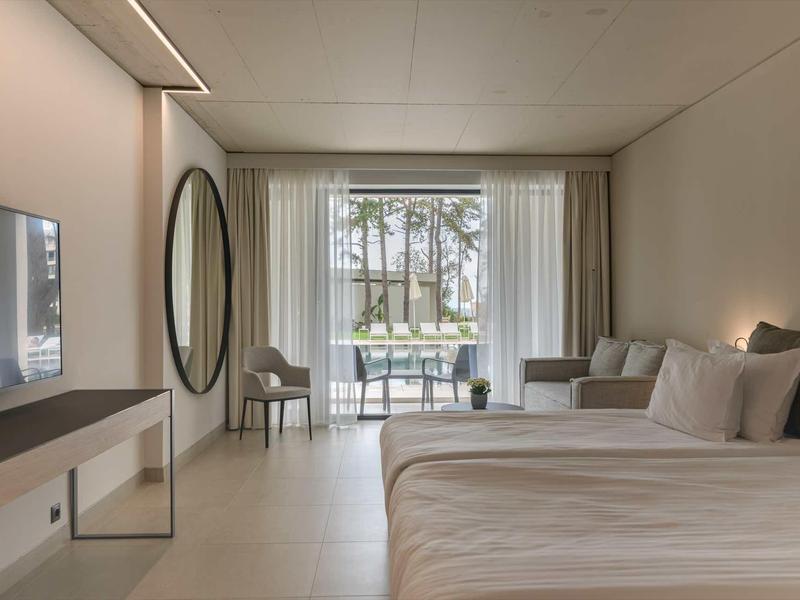 Modern hotel room with two beds, comfortable chairs, and a view of a balcony with garden.
