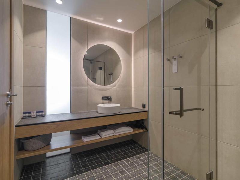 Modern bathroom with round mirror, wooden shelf, and walk-in shower with glass partition.