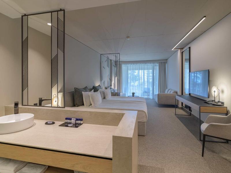 Modern hotel room with double bed, seating area, and large window with curtains.