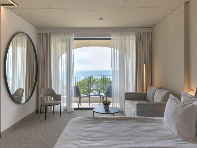 Bright hotel room with bed, sofa, chairs, and balcony overlooking the sea and trees.