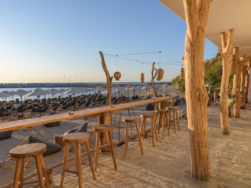 Open area with wooden stools and tables overlooking the beach and sea at sunset.