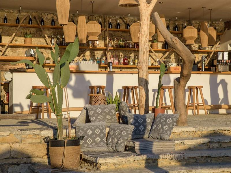 Outdoor lounge with cushions, plants, and bar stools in front of a bar at evening light.