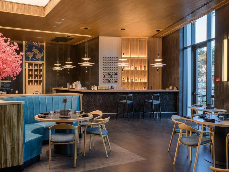 Modern restaurant with blue booths, wooden tables, and bar area under warm lighting.