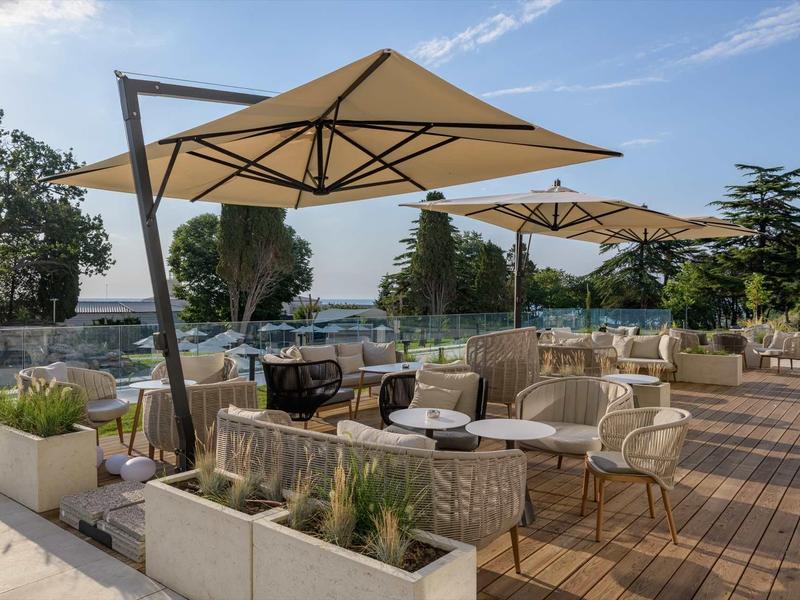 Sunny outdoor terrace with chairs, tables, and large umbrellas by a pool.