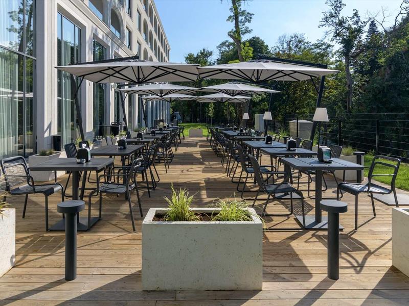 Terrace with tables and umbrellas next to a hotel building in a green garden.