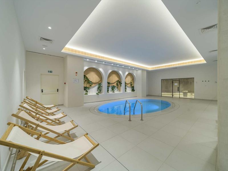 Modern indoor pool with round shape, deck chairs, and indirect ceiling lighting.