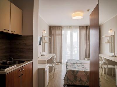 Kleines Hotelzimmer mit Küche, Schreibtisch, Bett und großem Fenster mit Vorhängen.