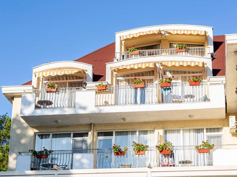 Weißes Haus mit zwei Balkonen, blauen Himmel, Blumen und roten Ziegeldächern.