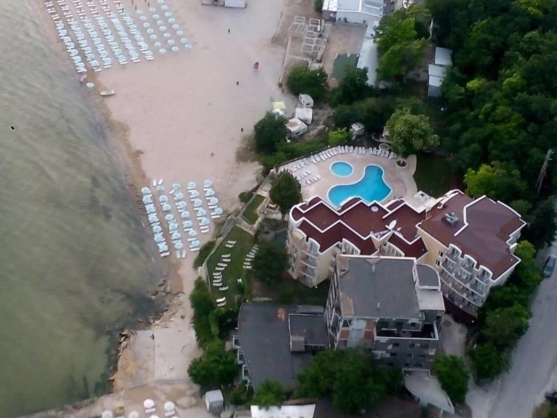 Luftaufnahme eines Resorts mit Pool, Strand und Liegestühlen an bewaldeter Küste.