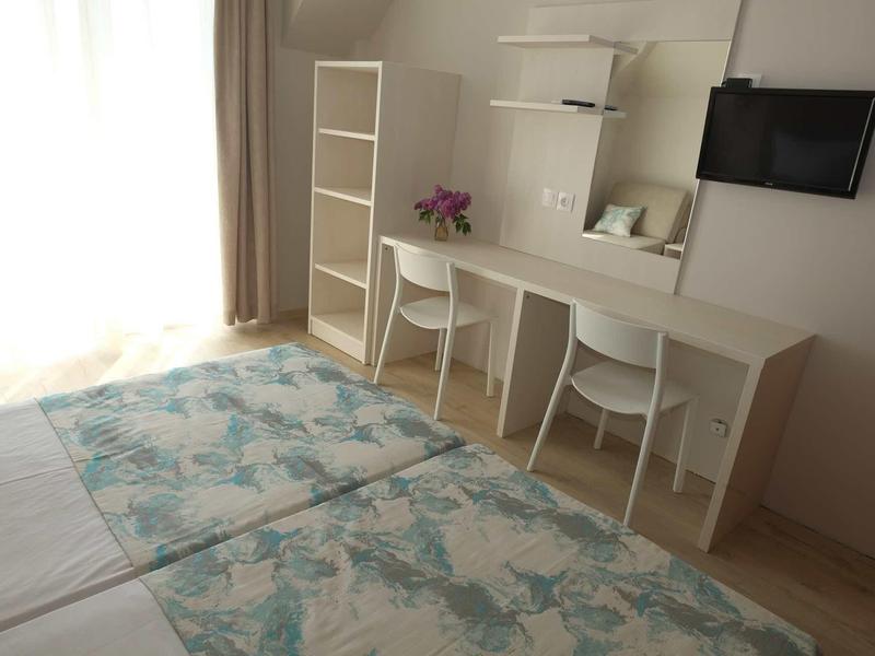 Helles Zimmer mit zwei Betten, Schreibtisch, weißen Stühlen, Regal und Fernseher an der Wand.