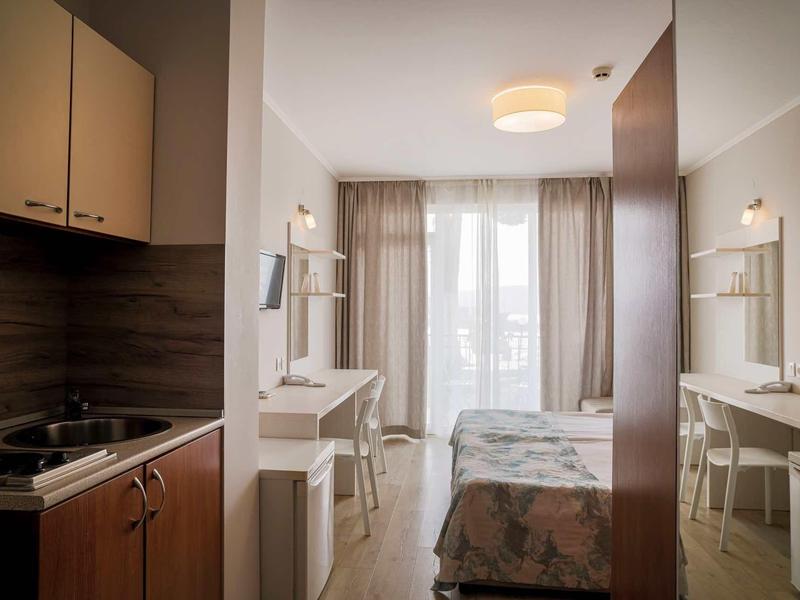 Kleines Hotelzimmer mit Küche, Schreibtisch, Bett und großem Fenster mit Vorhängen.