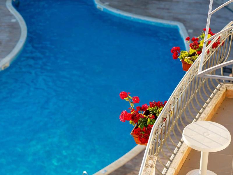Blauer Pool mit klaren Wasser, daneben Balkon mit weißen Stühlen und roten Blumentöpfen.