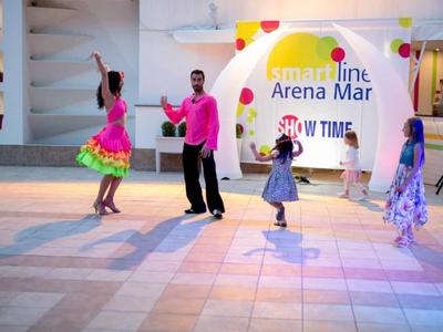 Taneční pár v barevném oblečení předvádí představení pro dvě malé diváky u značky Arena Mar.