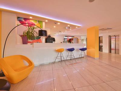 Lichte hotelreceptie met witte balie, drie kleurrijke barkrukken en een oranje fauteuil.