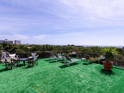 Dachterrasse mit grünem Boden, Liegestühlen, Tischen und Pflanzen mit Blick auf Bäume und Himmel.