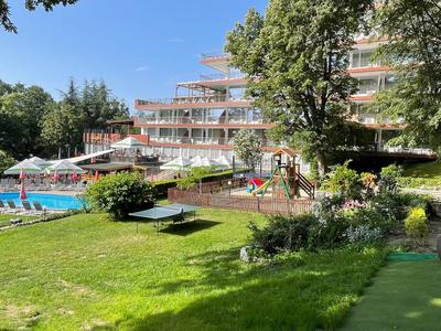 Bâtiment d'hôtel avec piscine extérieure, chaises longues et jardin vert par temps ensoleillé.