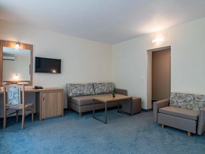 Hotelzimmer mit blauem Teppich, Sofa, Sessel, Schreibtisch und Wandfernseher.