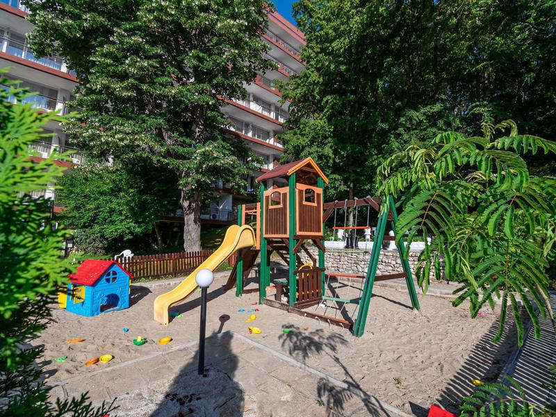 Kinderspielplatz mit Rutsche, Schaukeln und Sandkasten vor einem Wohngebäude und viel Grün.
