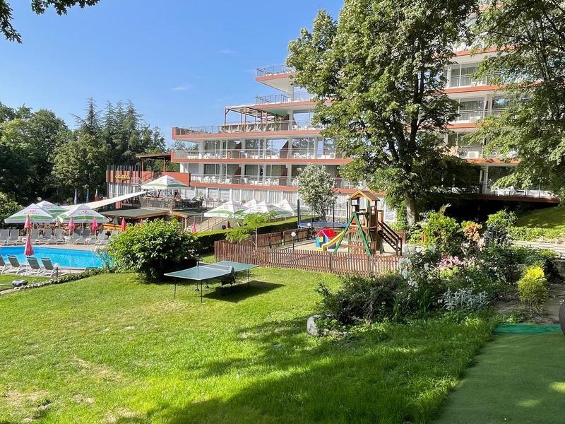 Hotel mit Außenpool, Liegestühlen und Sonnenschirmen in einer grünen Gartenanlage bei Sonnenschein.