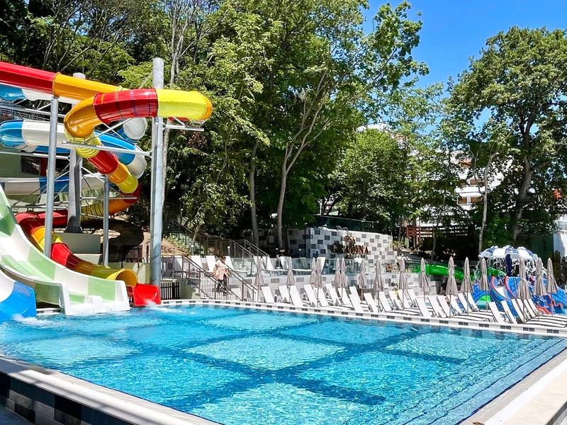 Ein Außenpool mit Wasserrutschen und Liegestühlen, umgeben von Bäumen an einem sonnigen Tag.