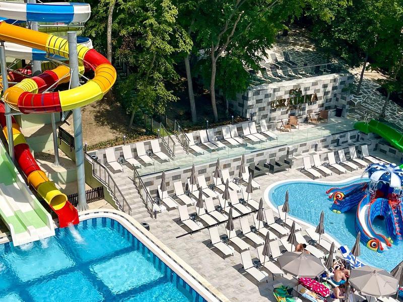 Wasserpark mit Rutschen und zwei Pools, umgeben von vielen Liegestühlen und Bäumen.