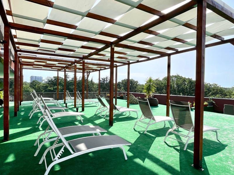 Sonnenterrasse mit Liegestühlen und Glasdach, umgeben von Bäumen und Blick auf die Stadt.