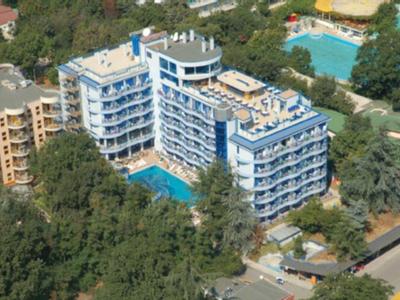 Hotel wielopiętrowy z dwoma basenami, otoczony zielonymi drzewami i innymi budynkami.