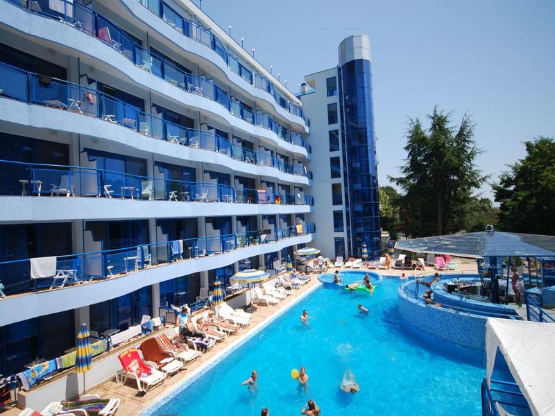 Wielopiętrowy hotel z niebieskim odkrytym basenem i leżakami otoczony drzewami.