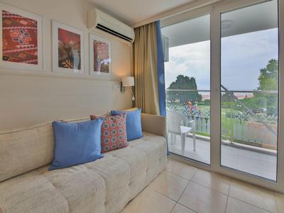 Jasny salon z sofą, kolorowymi poduszkami i dużymi szklanymi oknami z widokiem na przyrodę.