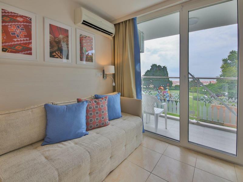 Jasny salon z sofą, kolorowymi poduszkami i dużymi szklanymi oknami z widokiem na przyrodę.