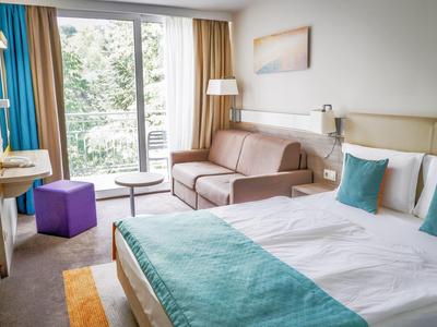 Nowoczesny pokój hotelowy z dużym łóżkiem, sofą, biurkiem i balkonem z widokiem na zieleń.