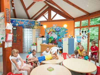 Kolorowe wnętrze przedszkola z kilkorgiem dzieci siedzących przy stołach i krzesłach.